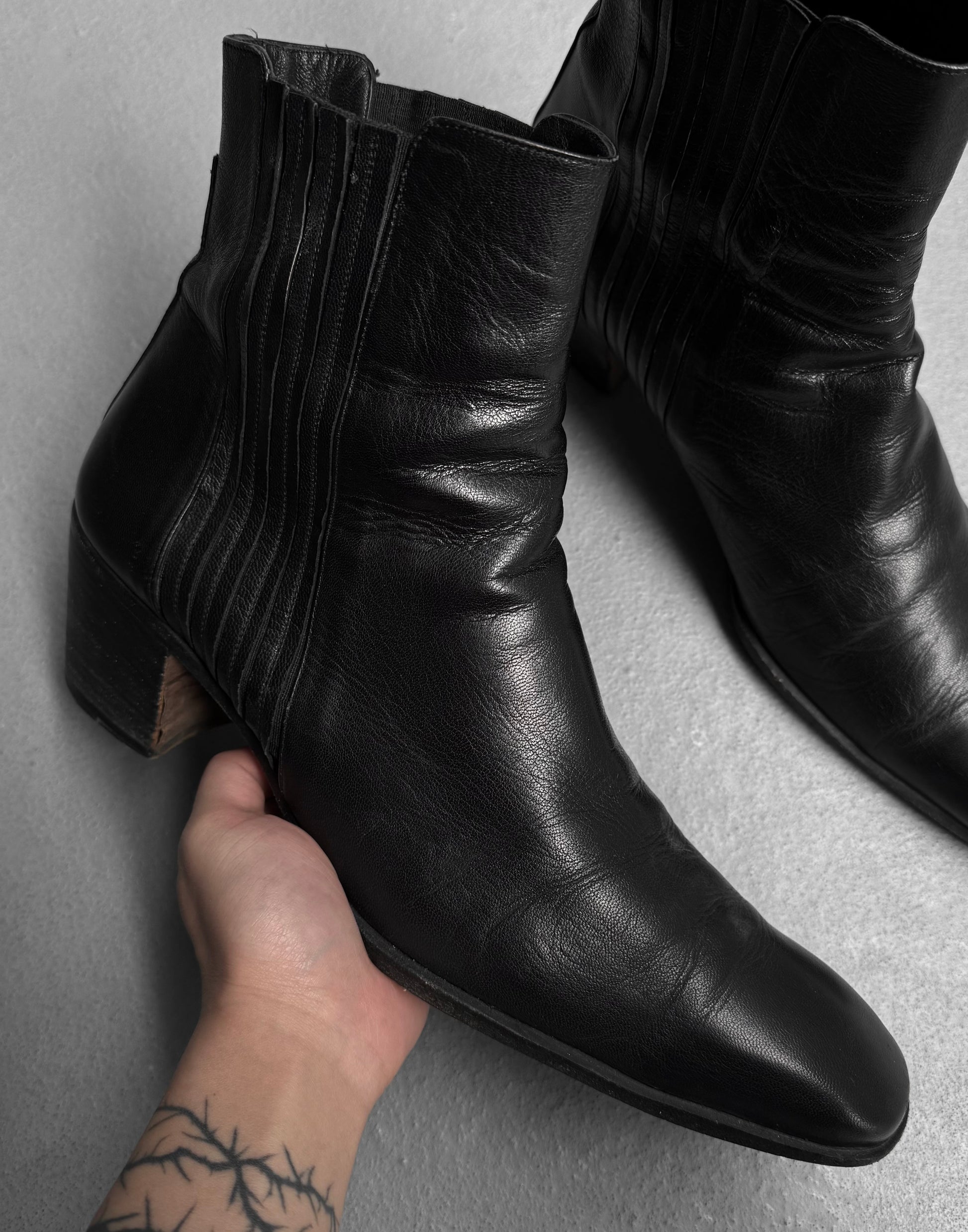 A hand with a barbed wire tattoo on the wrist holds a Saint Laurent Paris YSL Rive Gauche SS04 by Tom Ford 40mm Heel ‘Johnny’ Leather Boot, while another matching boot rests nearby on a light gray surface.