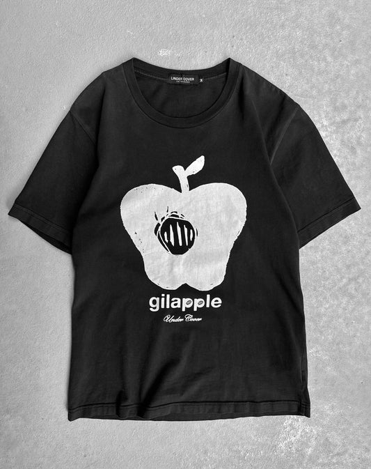 The Undercover SS16 ‘Gilapple’ Graphic Printed T-shirt features a large white apple and insect graphic with “gilapple” and UNDERCOVER text in white, showcasing Jun Takahashi’s streetwear style. The black shirt is displayed flat on a gray surface.