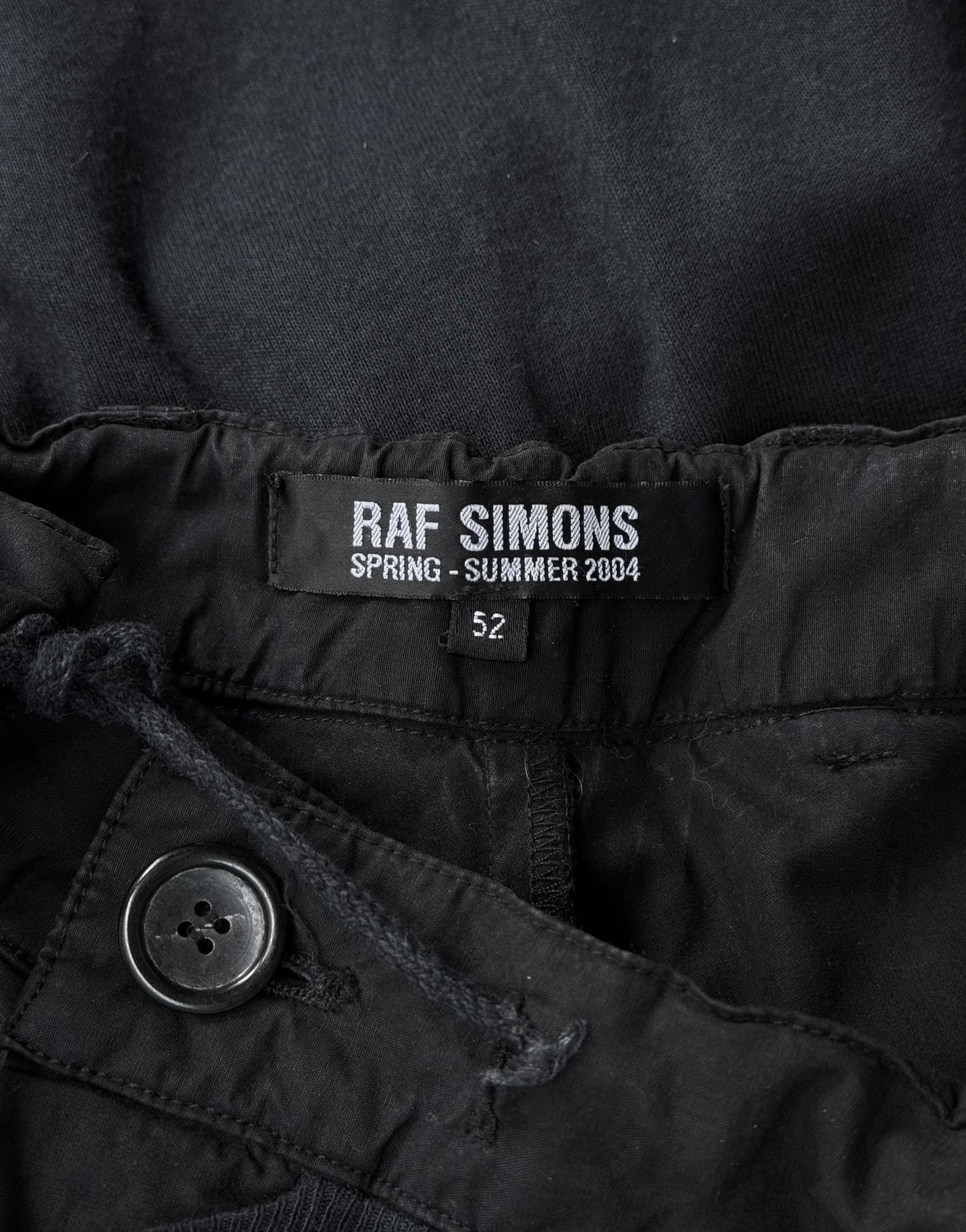 Close-up of a black label inside RAF SIMONS SS04 May the Circle Be Unbroken Reconstructed Zombie Wide Leg Pants, reading RAF SIMONS SPRING - SUMMER 2004 with size “52” below. Button and drawstring add minimalistic detail.