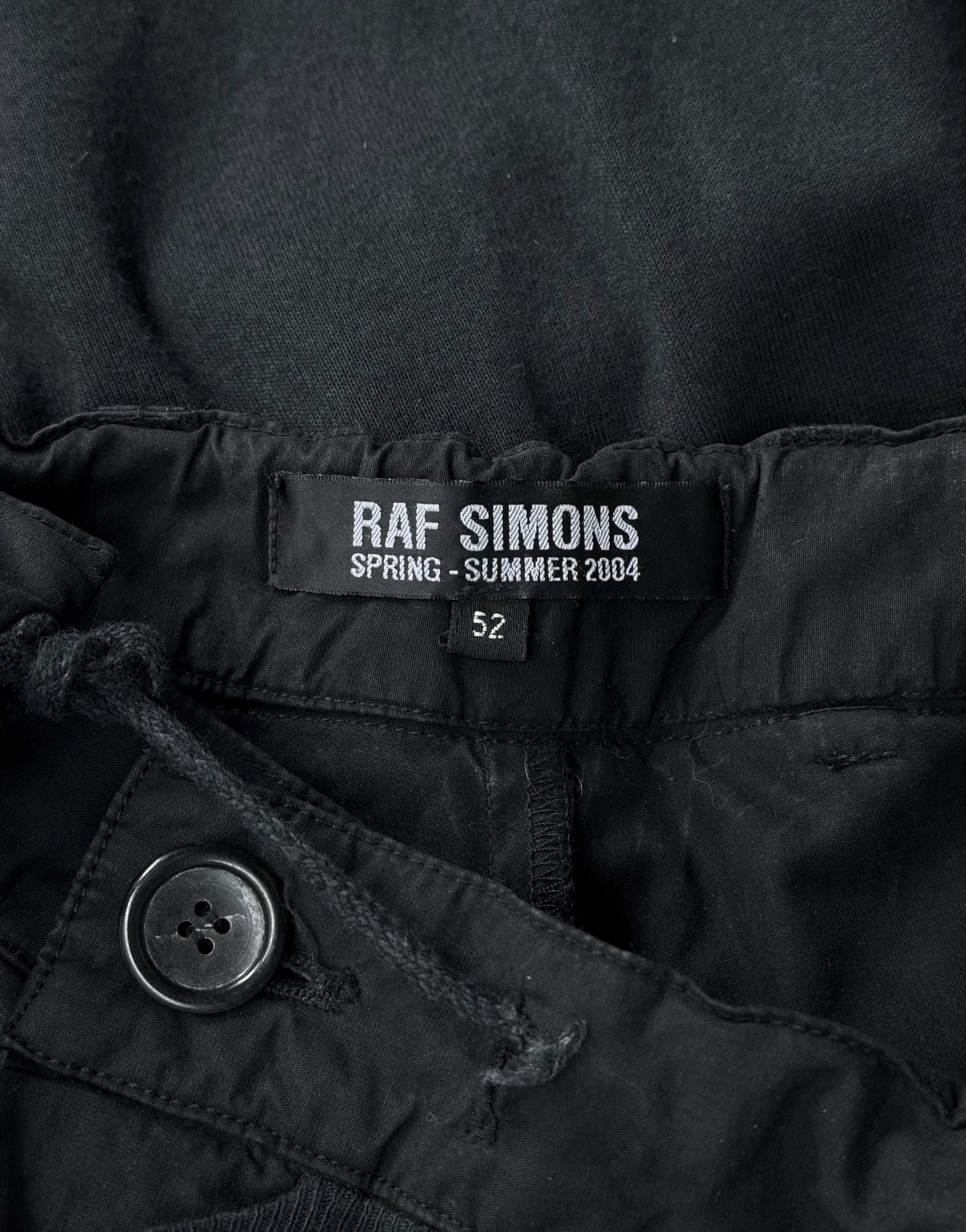 Close-up of a black label inside RAF SIMONS SS04 May the Circle Be Unbroken Reconstructed Zombie Wide Leg Pants, reading RAF SIMONS SPRING - SUMMER 2004 with size “52” below. Button and drawstring add minimalistic detail.