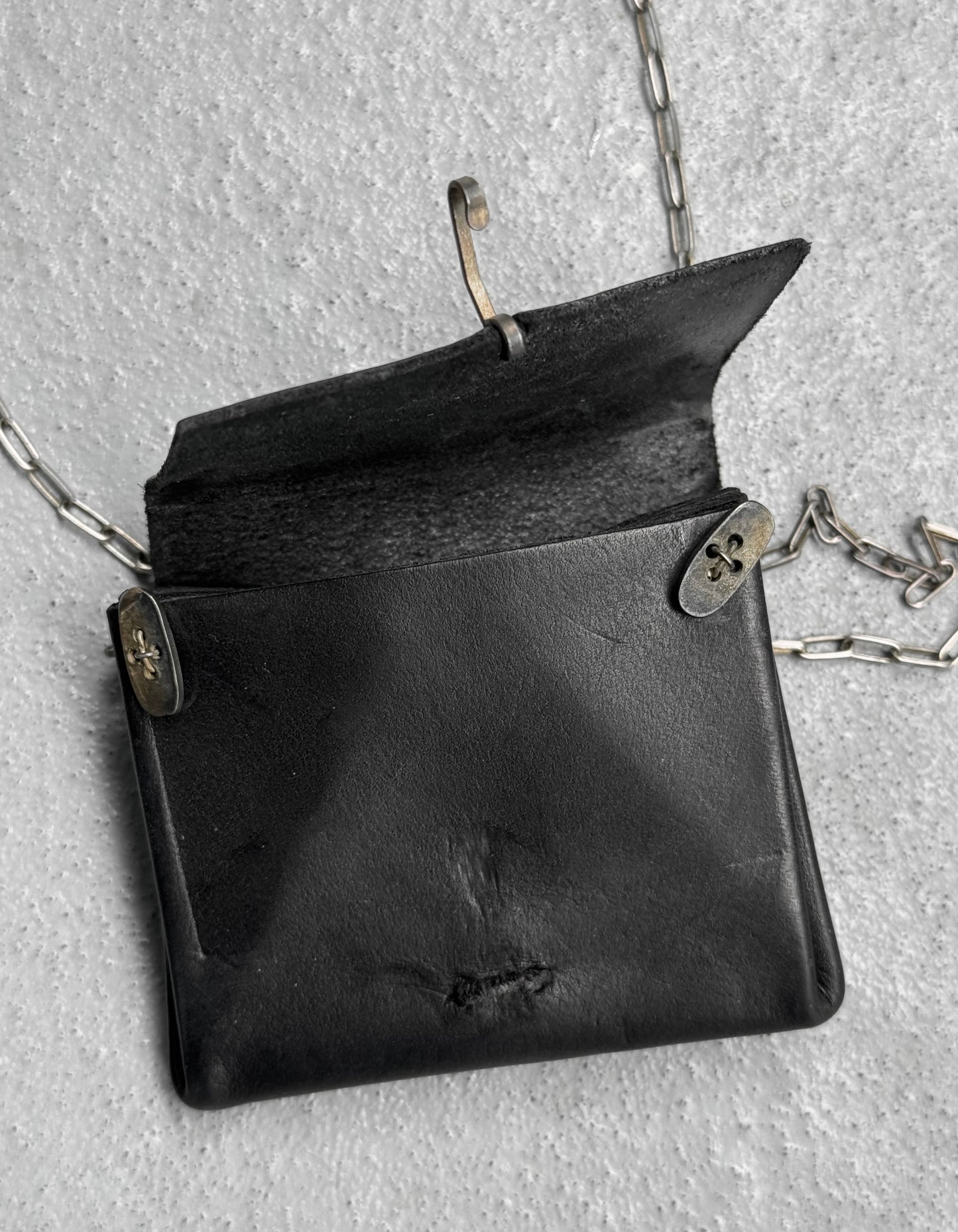 The M.A+ by Maurizio Amadei ‘A-B7201’ Calfskin Leather Mini Accordion Pouch Necklace features black leather, metal clasps, a silver chain, and an open flap, resting stylishly on a textured gray surface. OneSize holds your essentials.