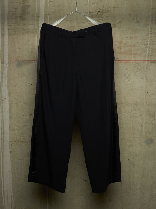 Black wide-leg cropped trousers hanging on a metal hanger against a textured concrete wall.