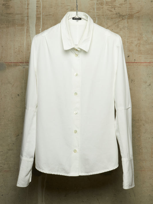 A white long-sleeve button-up shirt with a pointed collar, hanging on a black hanger against a textured concrete wall.