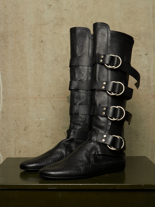 A pair of tall black leather boots with multiple decorative straps and large silver buckle details, displayed on a dark surface against a textured beige wall.