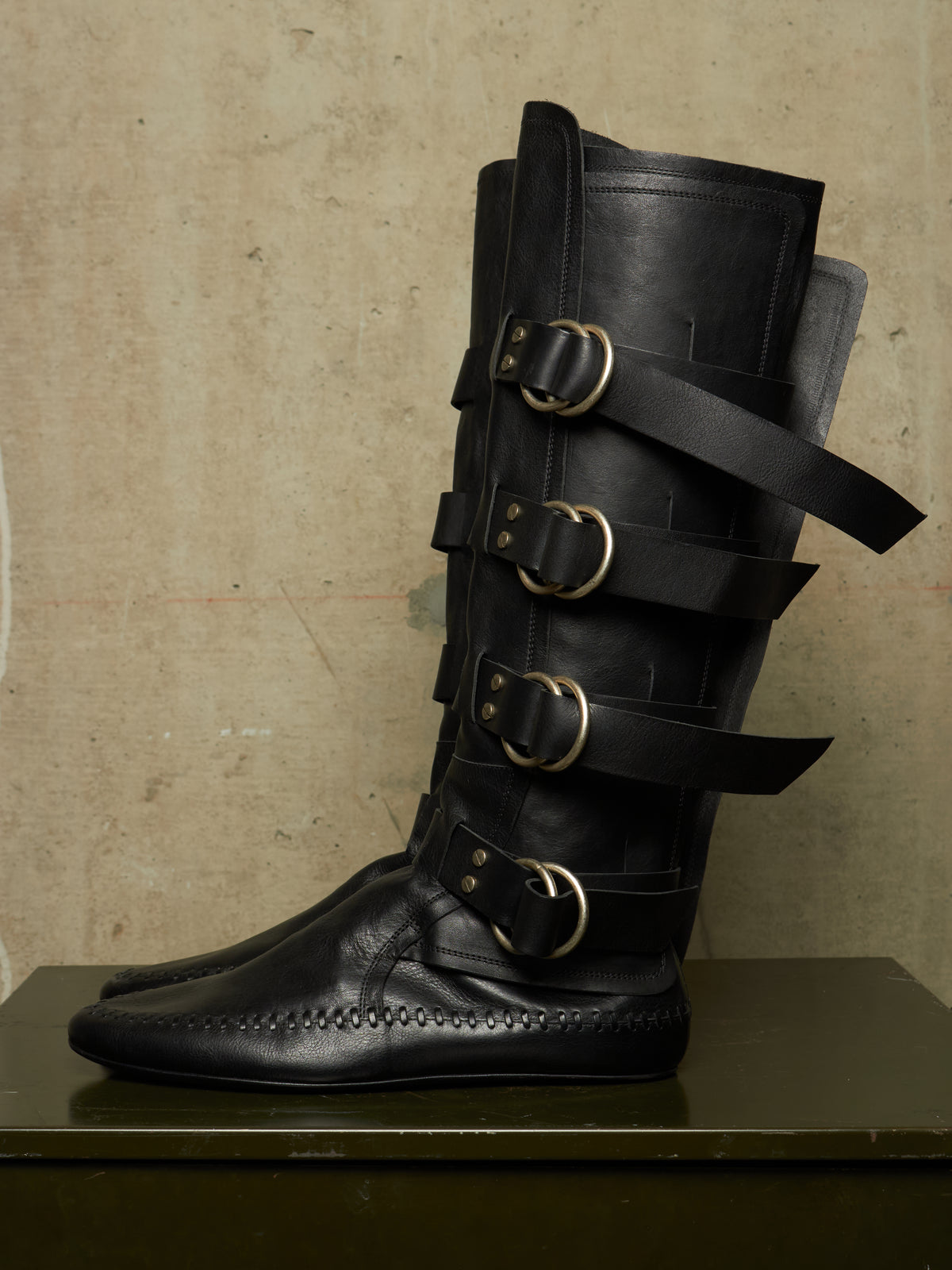 A pair of black leather knee-high boots with multiple decorative straps and large brass-tone buckles, displayed on a dark surface against a textured beige wall.
