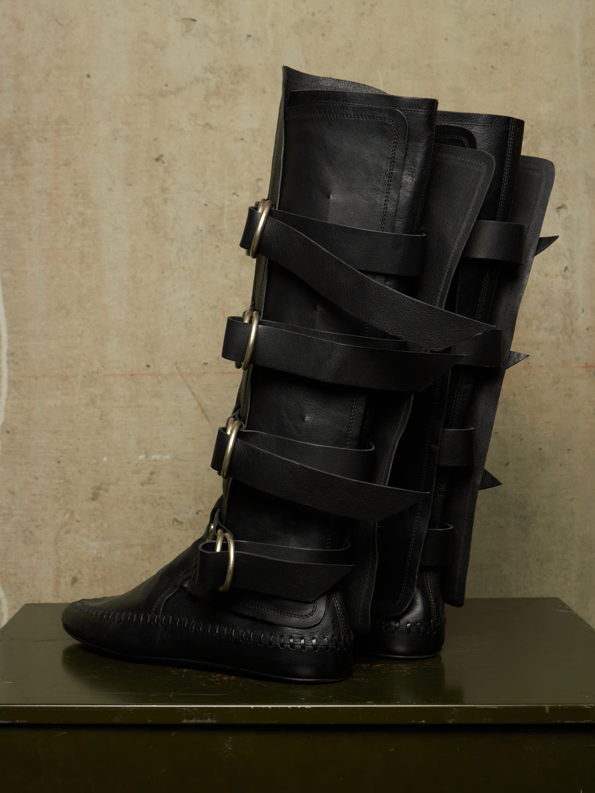 A pair of tall black leather boots with multiple wraparound straps and large metal buckles, displayed on a dark surface against a textured beige wall.