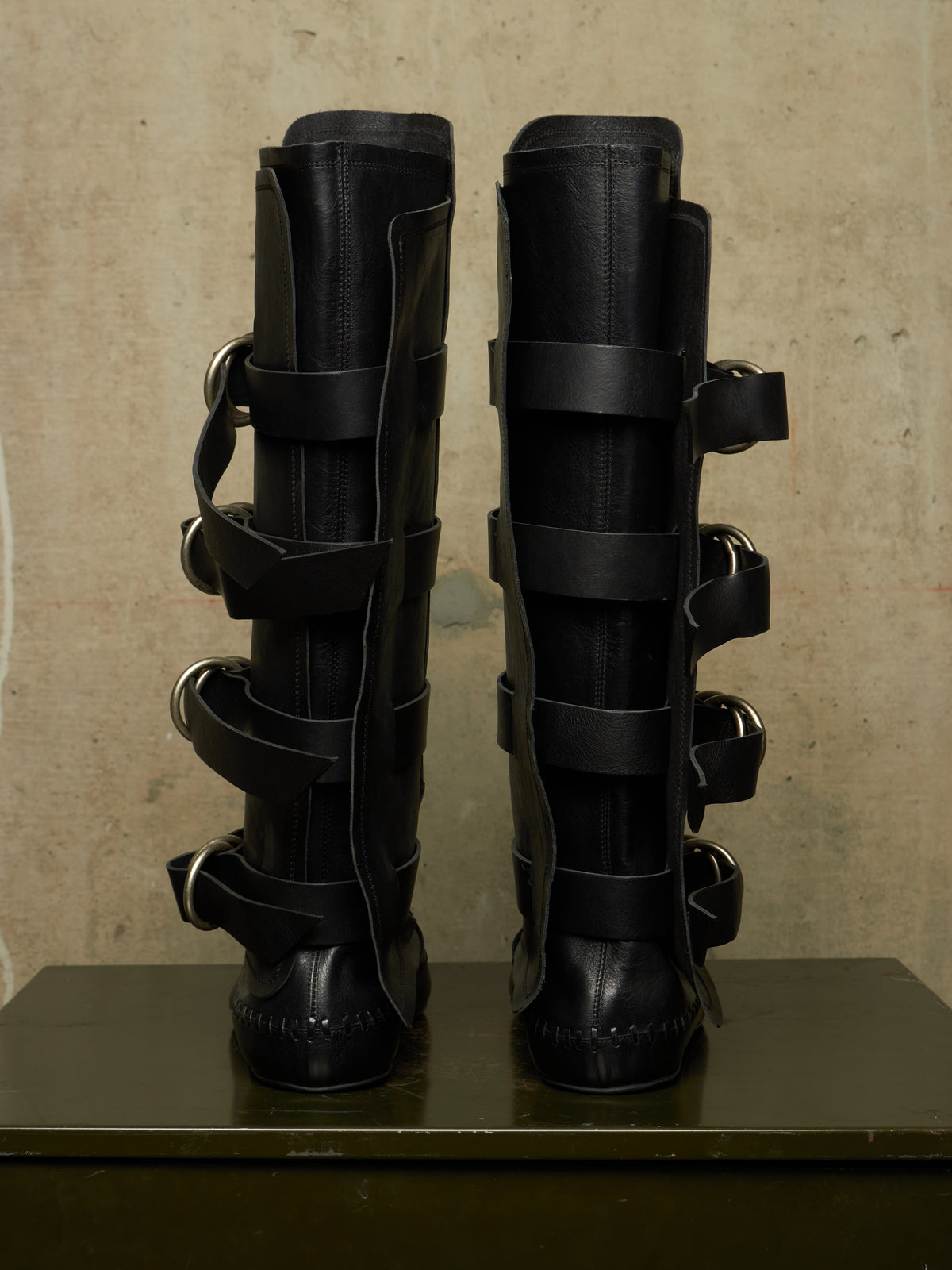 A pair of tall black leather boots with multiple wraparound straps and silver-tone metal rings, displayed upright on a dark surface against a textured beige wall.