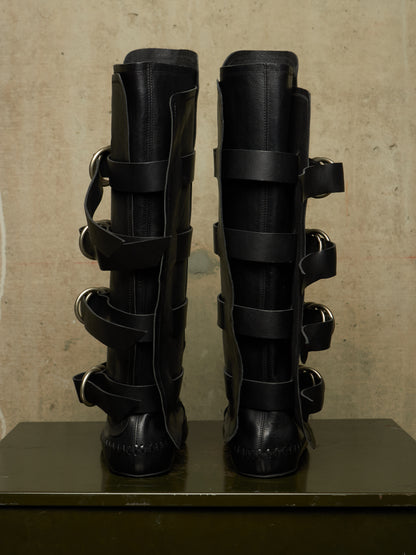 A pair of tall black leather boots with multiple wraparound straps and silver-tone metal rings, displayed upright on a dark surface against a textured beige wall.