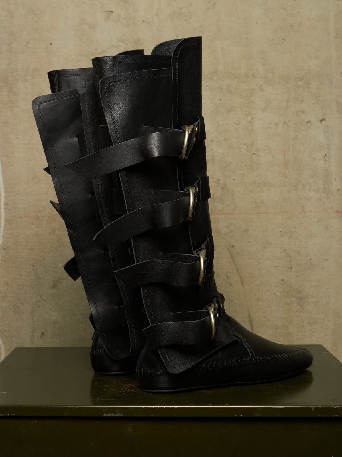 A pair of tall black leather boots with multiple wraparound straps and gold-tone buckles, displayed on a dark surface against a textured beige wall.