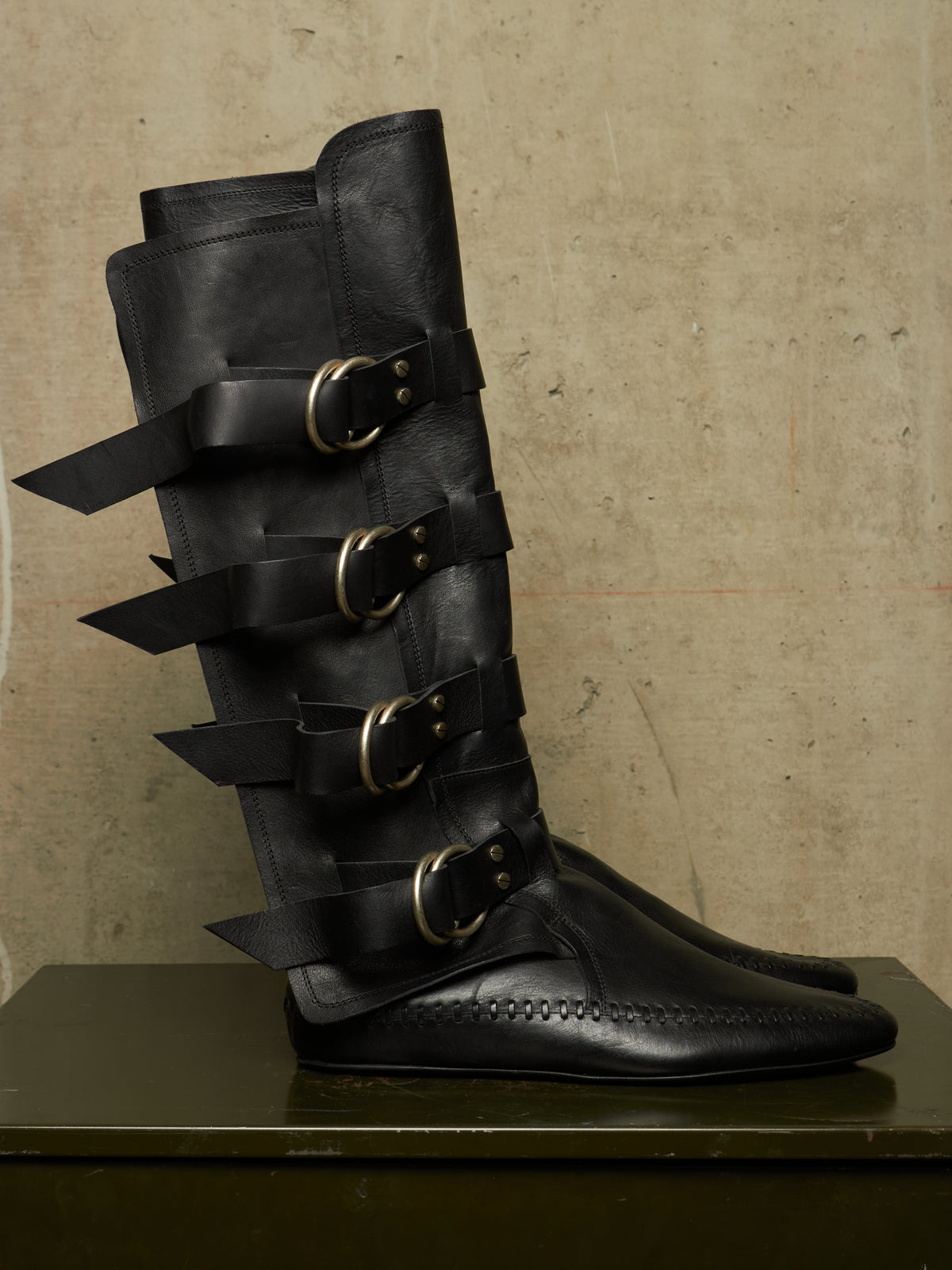 A tall black leather boot with multiple strap and buckle details, displayed on a dark surface against a textured beige wall.