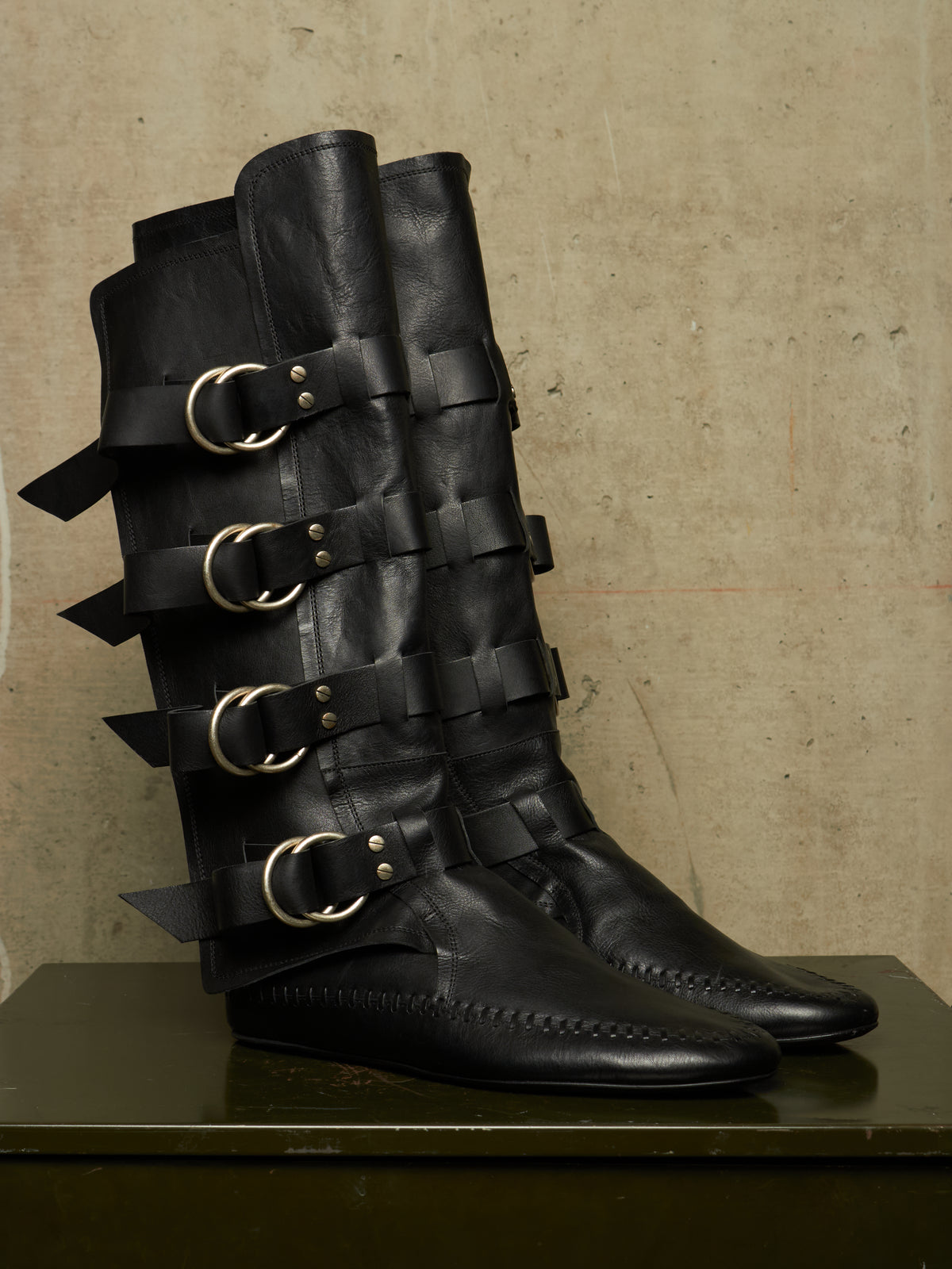 A pair of black leather knee-high boots with multiple decorative straps and large gold-tone buckle details, displayed on a dark surface against a textured beige wall.
