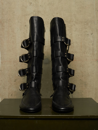 A pair of tall black leather boots with multiple wraparound straps and silver-tone metal buckles, displayed upright on a dark surface against a textured beige wall.