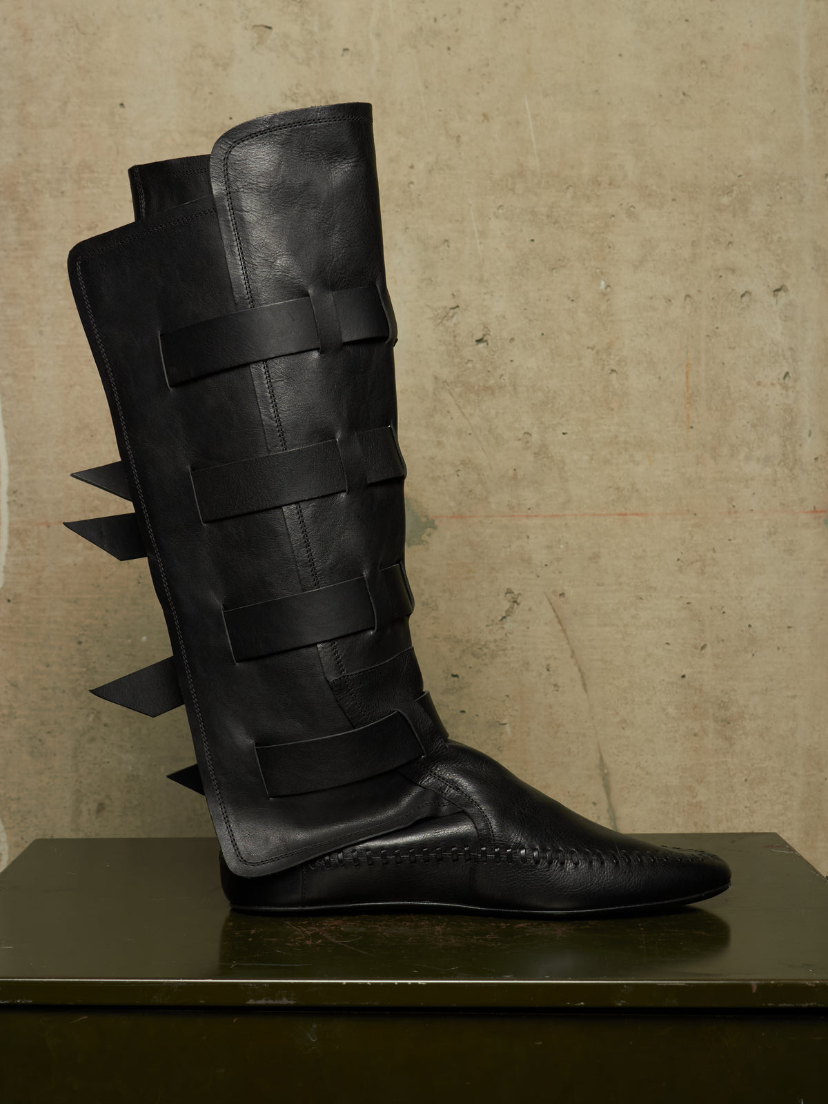 A single black leather knee-high boot with wide wraparound straps and sharp side spikes, displayed on a dark surface against a textured beige wall.