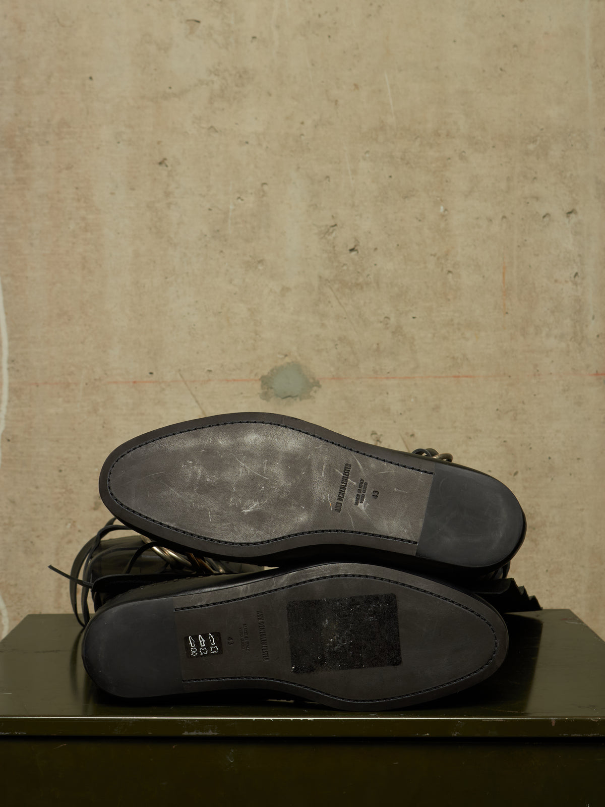 A pair of black leather shoes shown from the sole side, resting on a dark green surface against a textured beige wall.