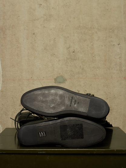 A pair of black leather shoes shown from the sole side, resting on a dark green surface against a textured beige wall.