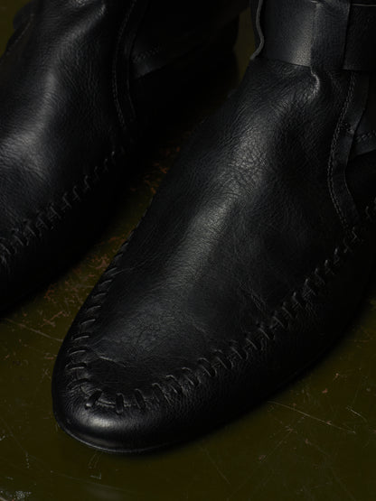 A close-up of a black leather moccasin boot with detailed stitching on a dark, textured olive-green surface.
