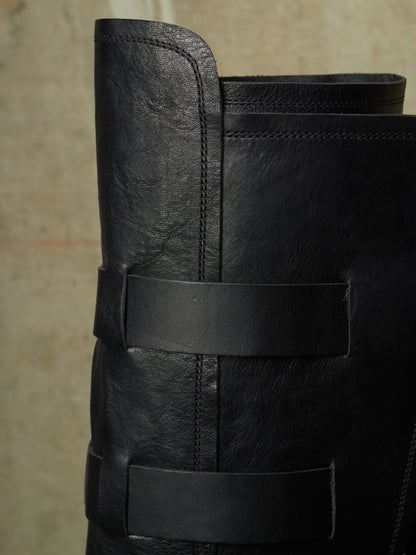 Close-up of a black leather boot with wide horizontal straps and visible stitching, set against a neutral textured background.