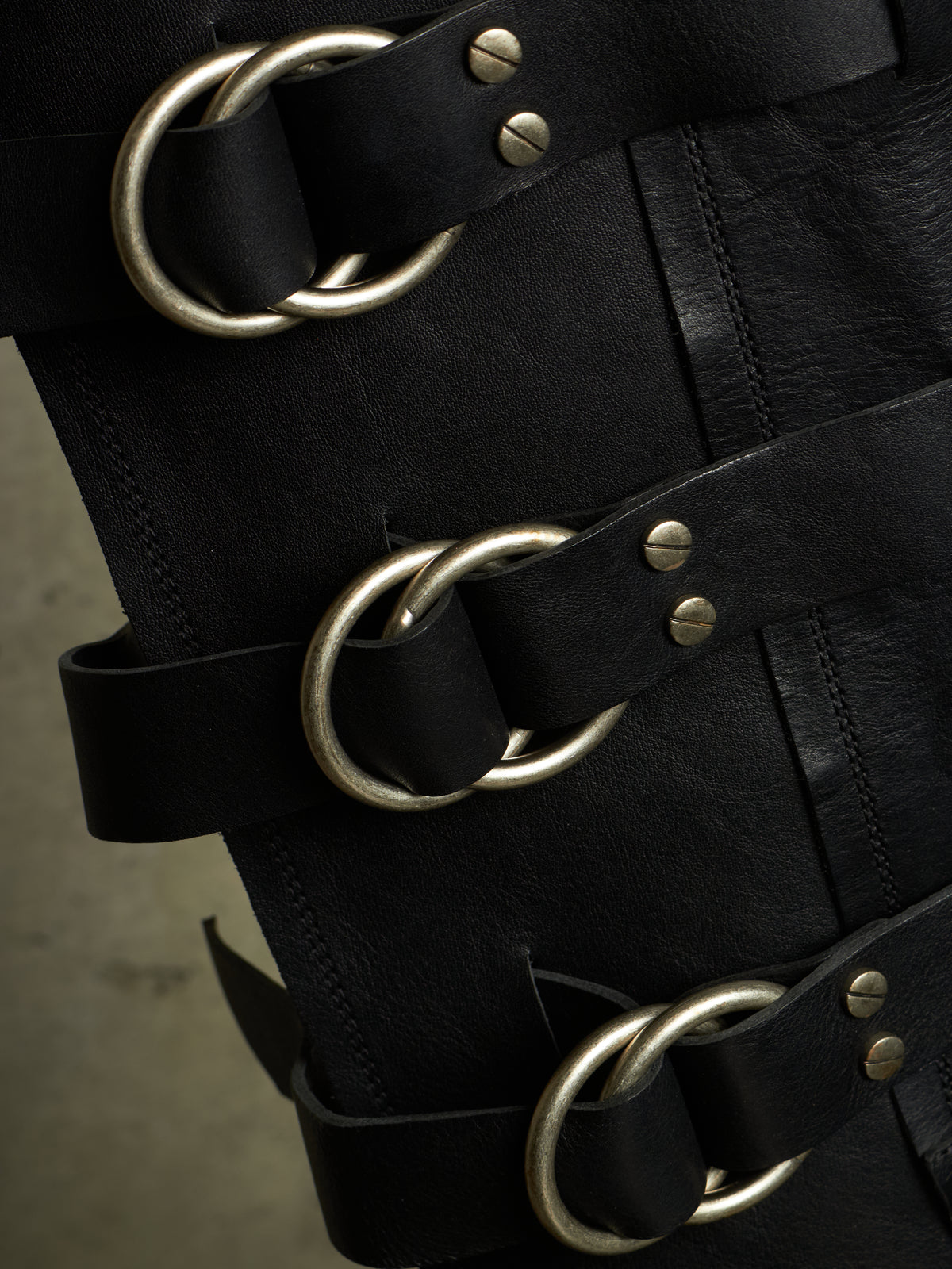 Close-up of a black leather strap accessory with large antique silver metal O-rings and rivets, shown against a dark background.