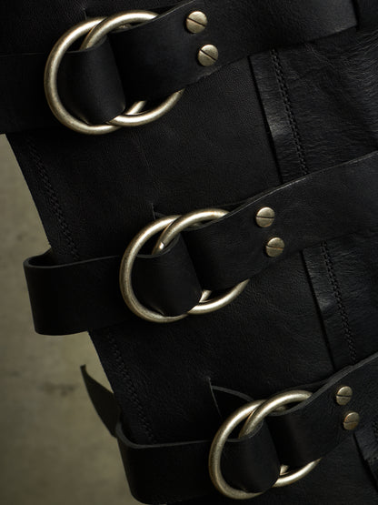 Close-up of a black leather strap accessory with large antique silver metal O-rings and rivets, shown against a dark background.