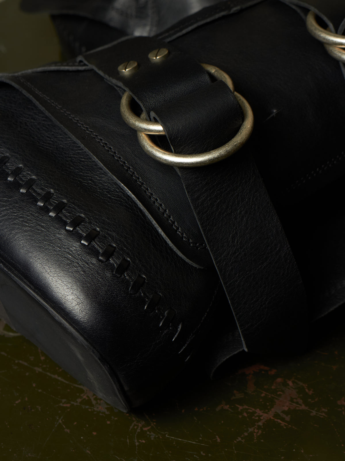 Close-up of a black leather bag with a wide strap, brass-tone ring hardware, and decorative stitching, resting on a dark textured surface.