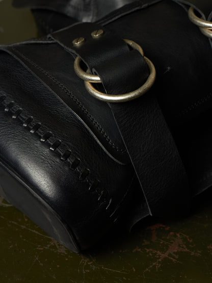 Close-up of a black leather bag with a wide strap, brass-tone ring hardware, and decorative stitching, resting on a dark textured surface.