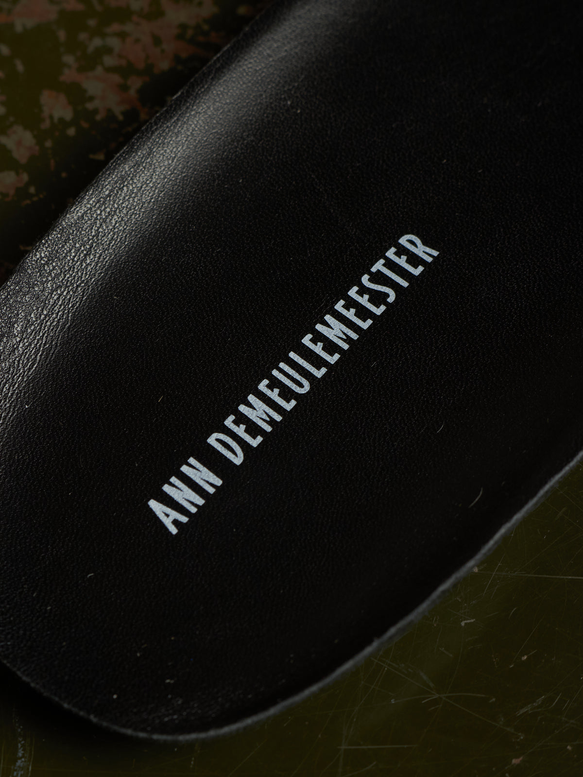 Close-up of a black leather shoe insole with the brand name 'ANN DEMEULEMEESTER' printed in white text, resting on a dark olive green textured surface.
