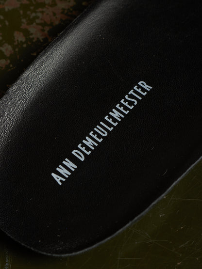 Close-up of a black leather shoe insole with the brand name 'ANN DEMEULEMEESTER' printed in white text, resting on a dark olive green textured surface.