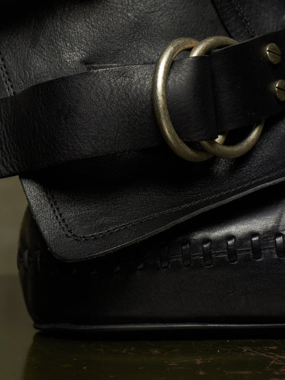 Close-up detail of a black leather bag with a wide strap featuring a large double round brass ring and a stitched edge.