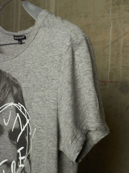 A close-up of a heather gray t-shirt hanging on a clear plastic hanger against a textured, warm-toned wall. The shirt features a partial graphic print of a person's face in black and white.
