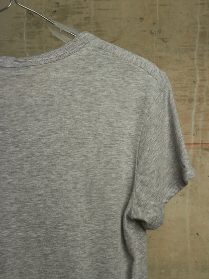 A close-up of a heather gray short-sleeve t-shirt hanging on a clear plastic hanger against a textured concrete wall.