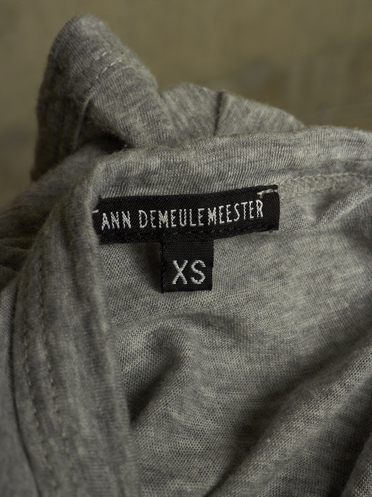 Close-up of a gray knit garment showing a black brand label that reads ANN DEMAULEMEESTER and a smaller size label reading XS.