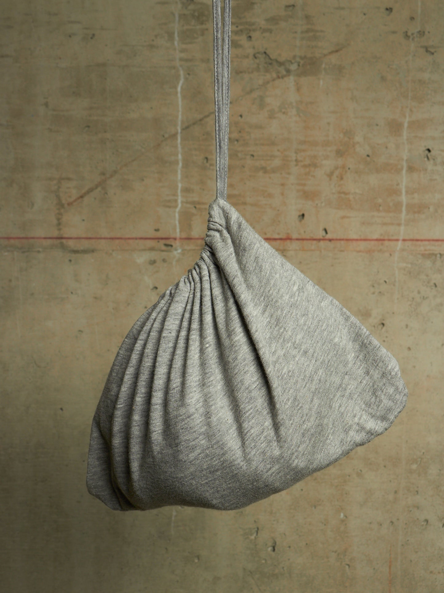 A light gray heathered fabric drawstring bag hanging from a matching cord against a textured concrete wall.