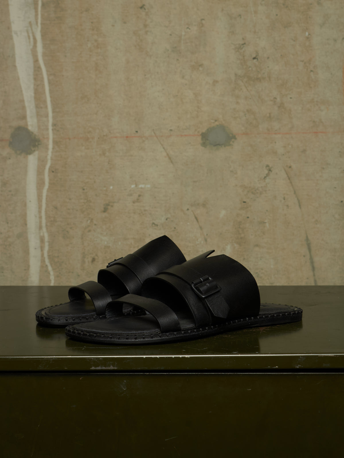 A pair of black leather double-strap slide sandals with buckle closures, displayed on a dark green surface against a textured beige concrete wall.