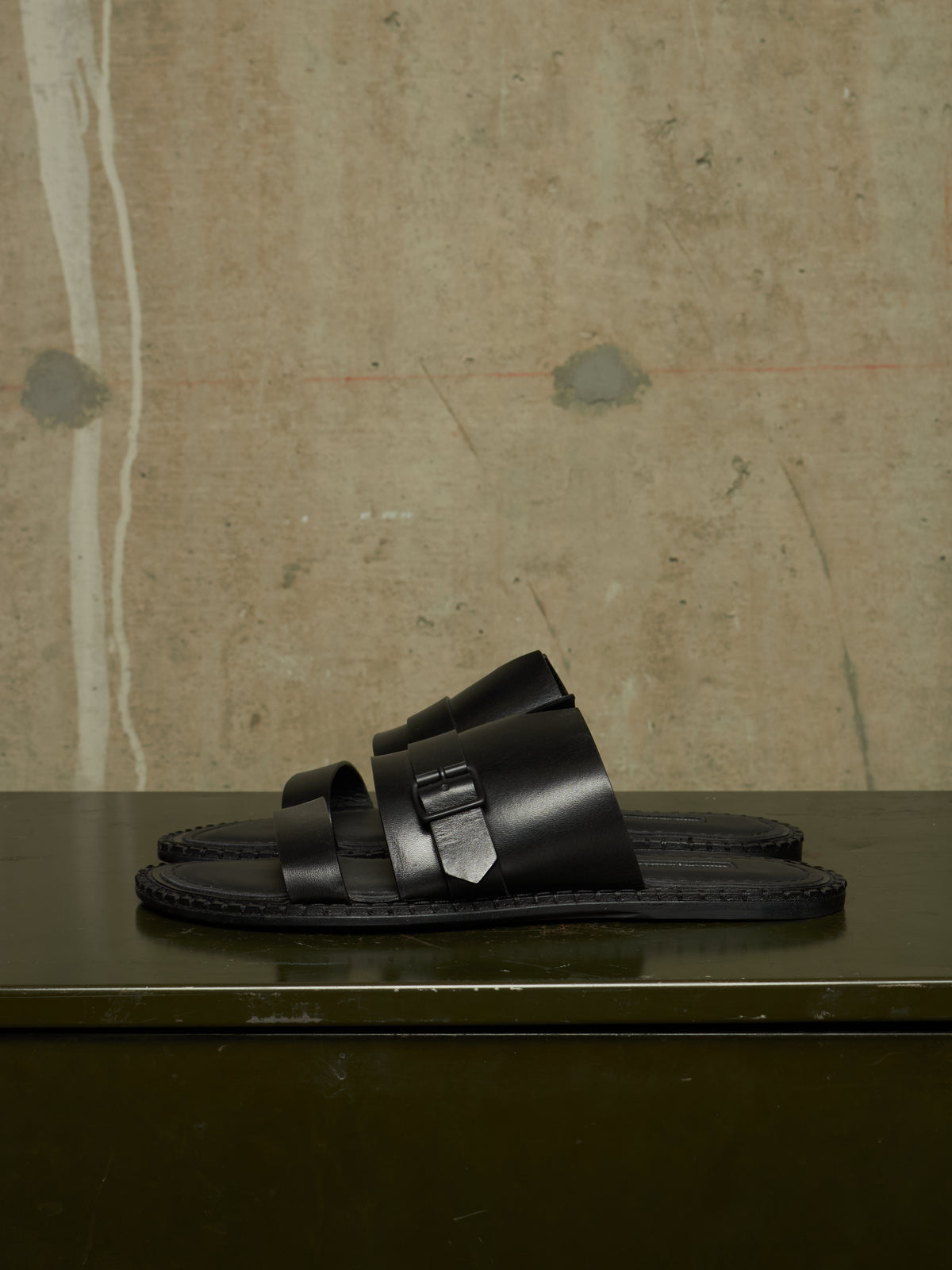 A pair of black leather double-strap slide sandals with a distinctive geometric metal buckle detail, displayed on a dark green surface against a textured concrete wall.