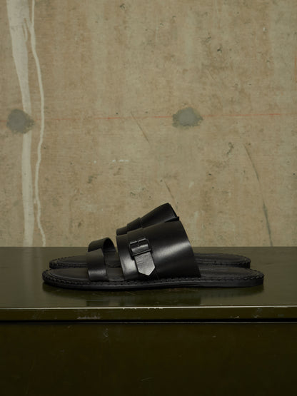 A pair of black leather double-strap slide sandals with a distinctive geometric metal buckle detail, displayed on a dark green surface against a textured concrete wall.