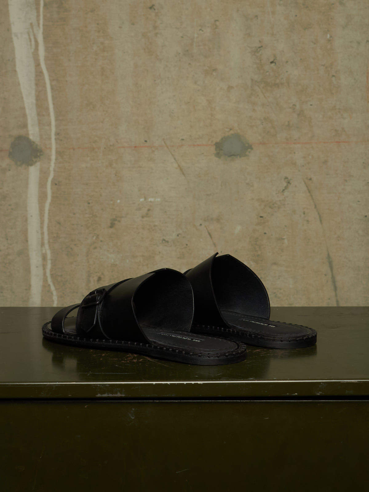 A pair of black leather slide sandals with wide straps and a buckle detail, resting on a dark green reflective surface against a textured beige concrete wall.