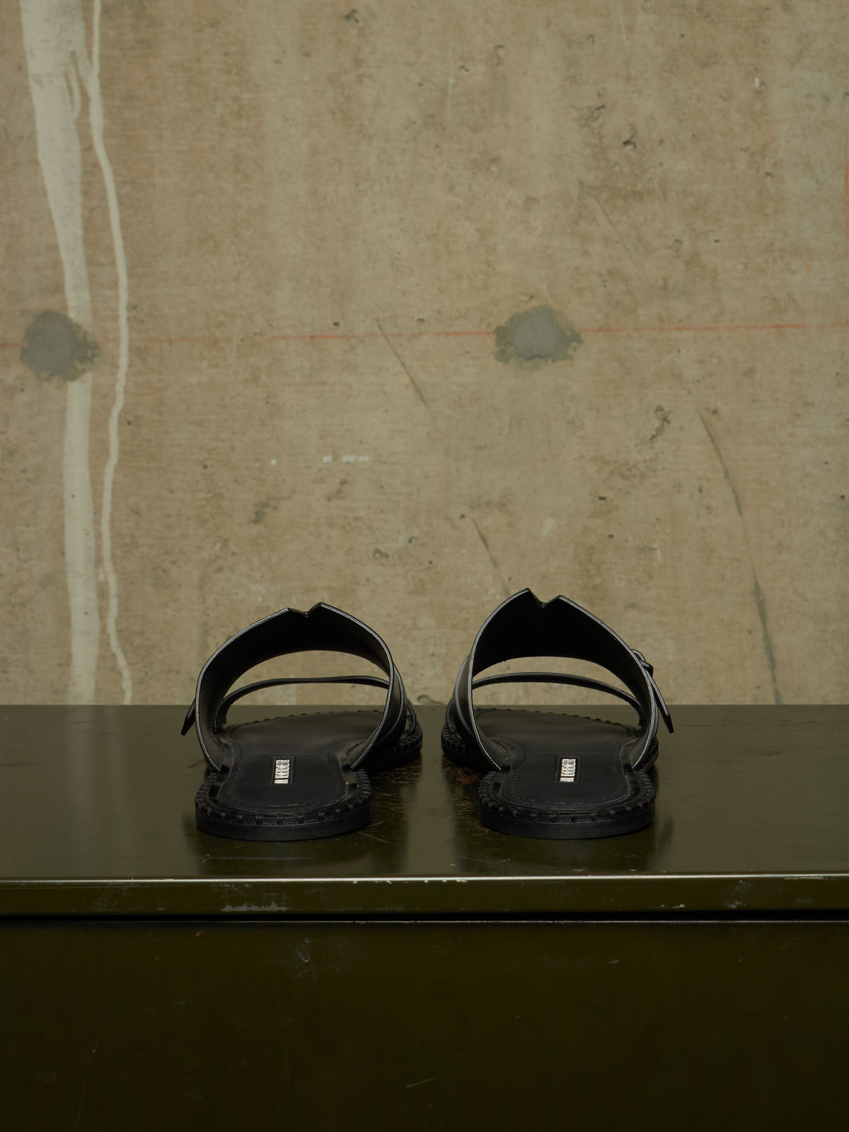 A pair of black leather slide sandals with double straps and a small silver rectangular logo plaque on the sole, displayed on a dark green surface against a textured beige concrete wall.