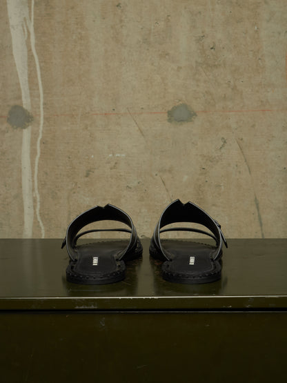 A pair of black leather slide sandals with double straps and a small silver rectangular logo plaque on the sole, displayed on a dark green surface against a textured beige concrete wall.