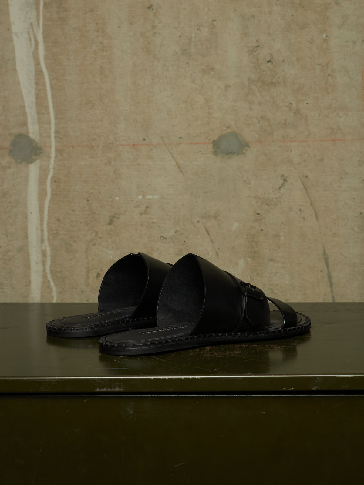 A pair of black leather slide sandals with wide straps and a small buckle detail, resting on a dark green surface against a textured concrete wall.