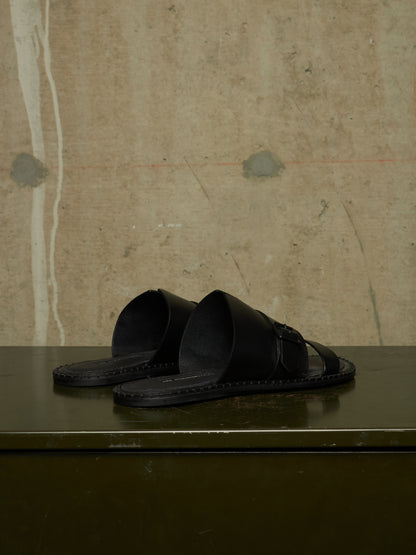 A pair of black leather slide sandals with wide straps and a small buckle detail, resting on a dark green surface against a textured concrete wall.