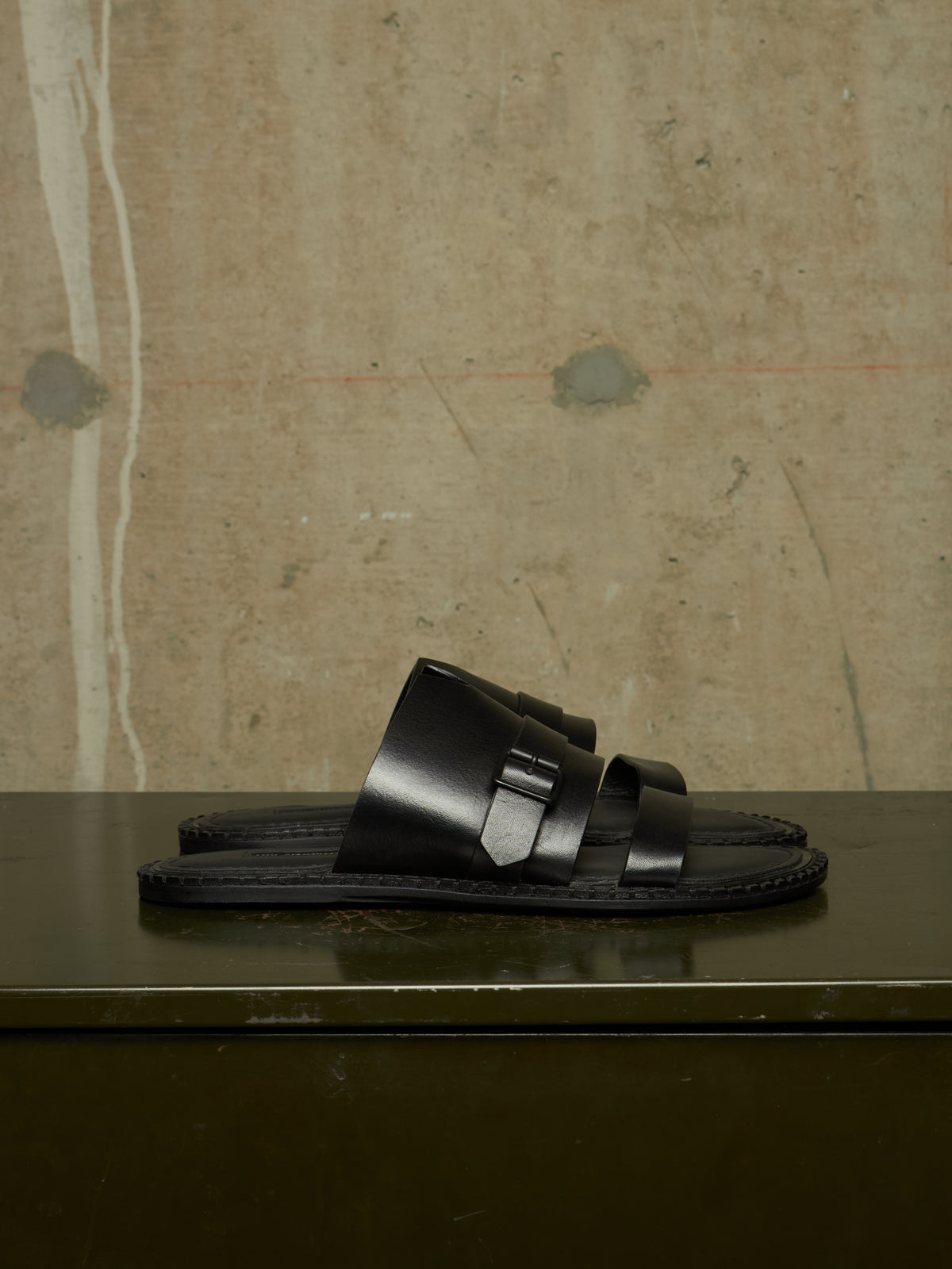 A pair of black leather double-strap slide sandals with silver buckle details, displayed on a dark reflective surface against a textured concrete wall.