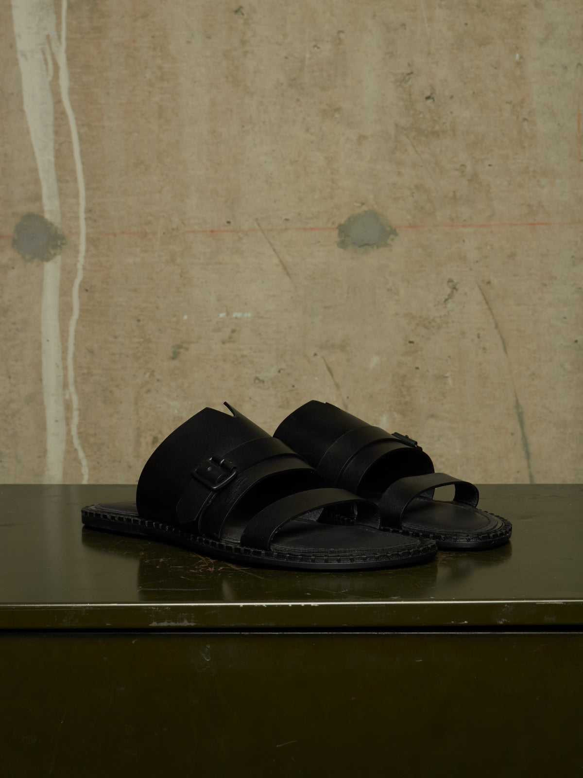 A pair of black leather slide sandals with double straps and buckle details, resting on a dark green table against a textured concrete wall.