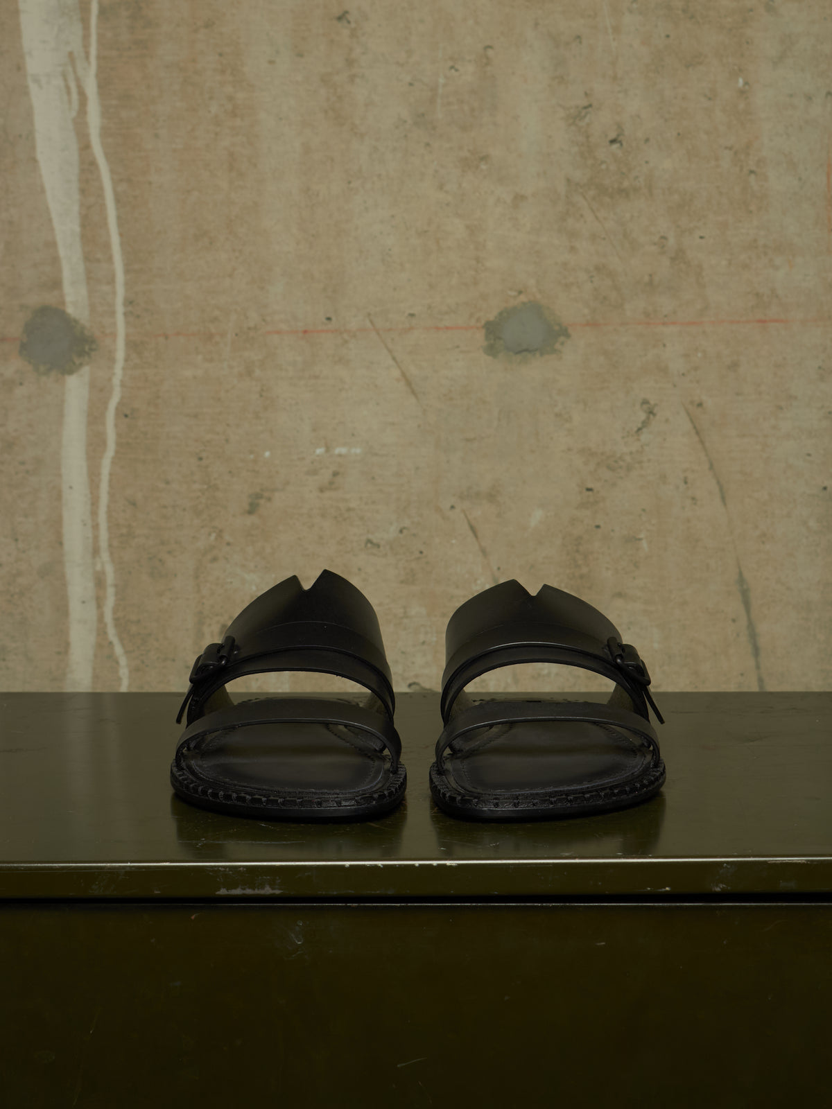 A pair of black leather slide sandals with wide straps and buckle details, displayed on a dark green surface against a textured beige concrete wall.
