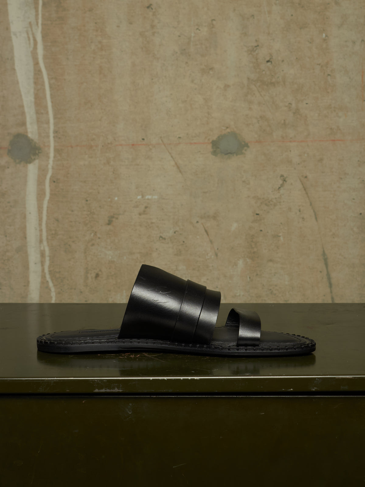 A single black leather slide sandal with wide stacked straps and a dark sole, displayed on a dark green surface against a textured beige wall.