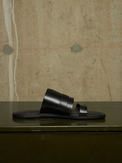 A single black leather slide sandal with wide stacked straps and a dark sole, displayed on a dark green surface against a textured beige wall.