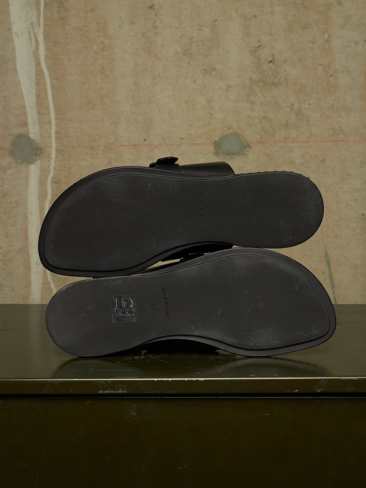A pair of black leather loafers shown from the bottom, displaying the outsoles and a small embossed logo, resting on a dark green surface against a textured beige wall.
