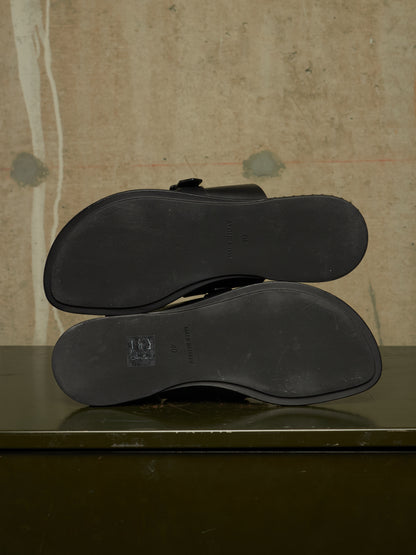 A pair of black leather loafers shown from the bottom, displaying the outsoles and a small embossed logo, resting on a dark green surface against a textured beige wall.