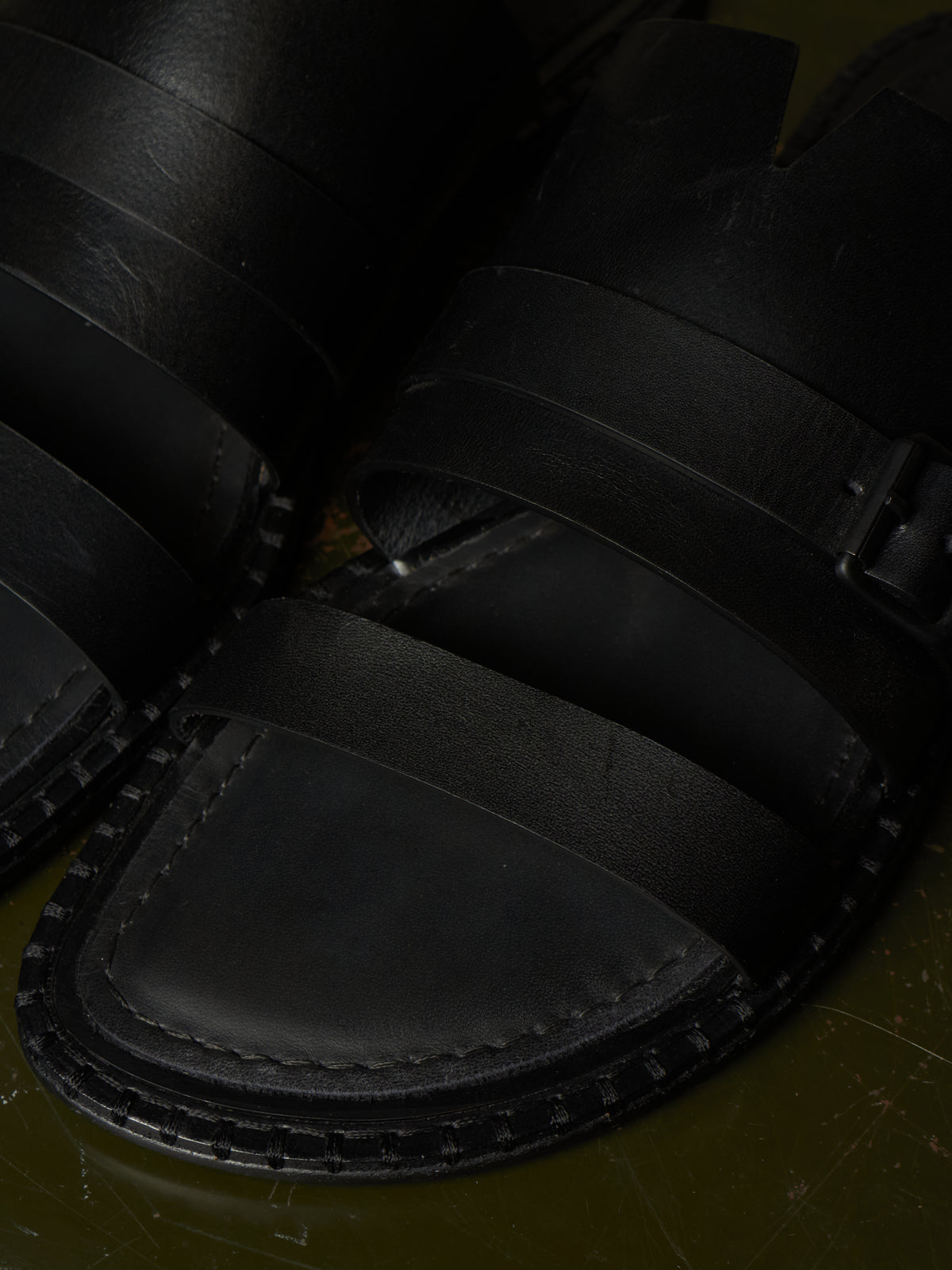 Close-up of black leather sandals with wide straps and a stitched sole, photographed against a dark olive-green surface.
