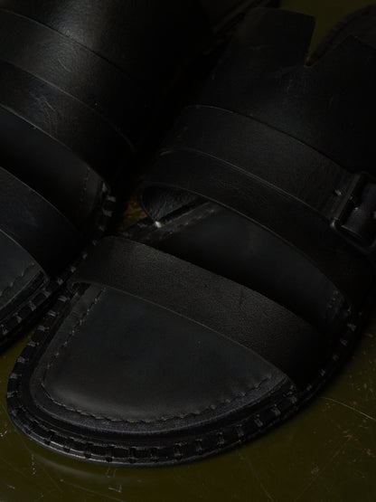 Close-up of black leather sandals with wide straps and a stitched sole, photographed against a dark olive-green surface.
