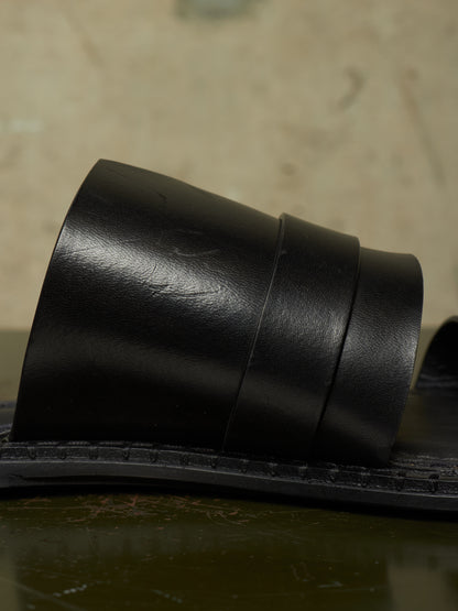 A close-up of a black leather slide sandal strap resting on a dark surface against a textured beige background.